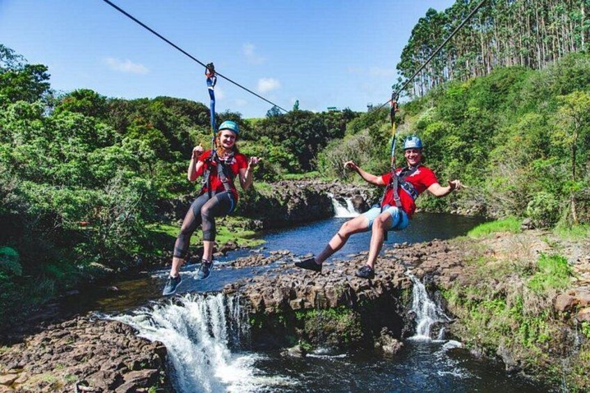 Two people zip-lining over a waterfall in a forested area. 1