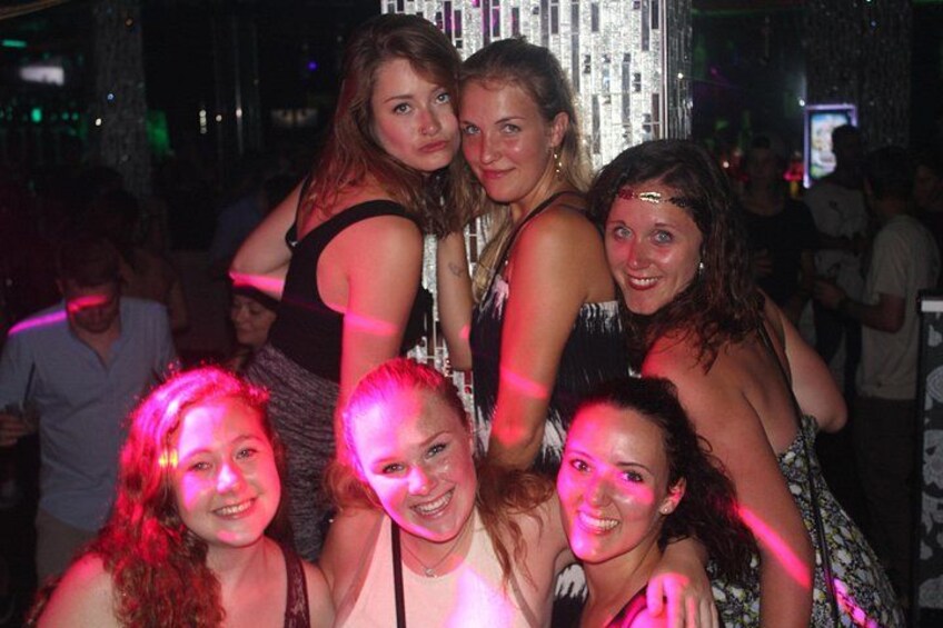 A group of people in a dimly lit indoor setting with reflective surfaces and a crowd in the background. 3