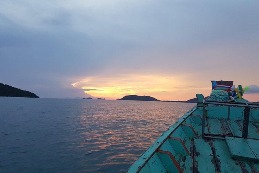 A boat with a blue and green color scheme, a person sitting at the back, and a sunset over the water with distant landforms. 1