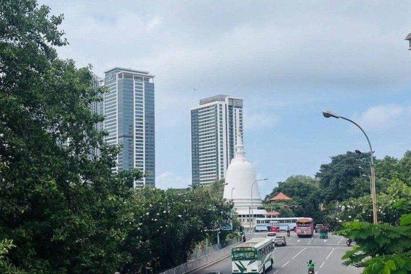 A city street with tall buildings, a white monument, and a green bus. 2