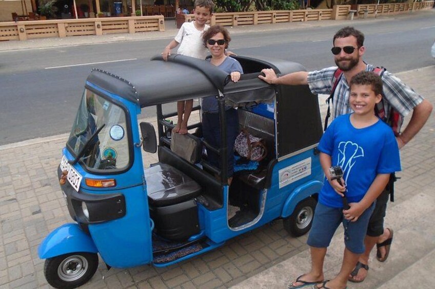 A blue auto-rickshaw parked on a sidewalk with three people standing beside it. 3