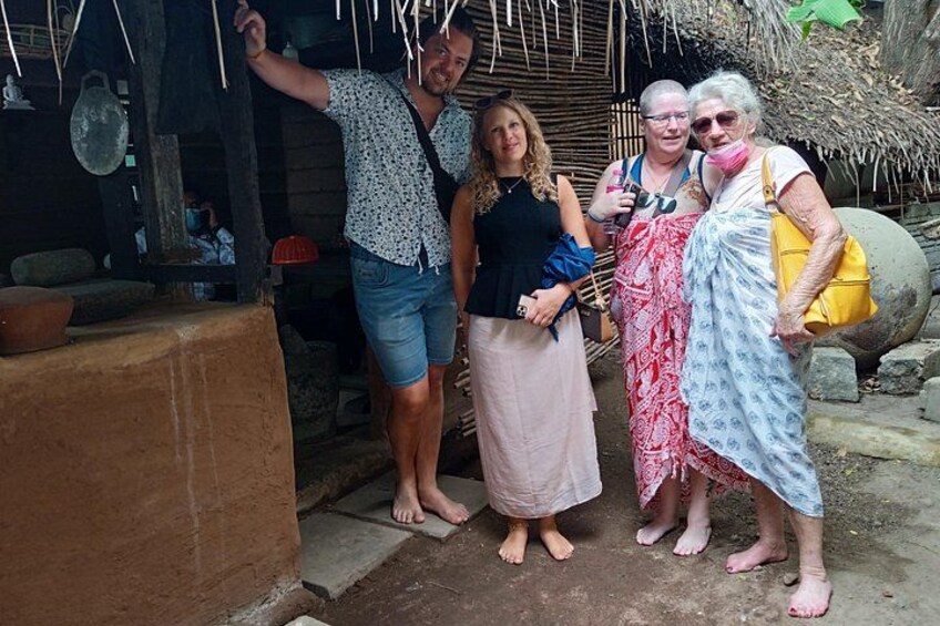 A group of people standing in a rustic outdoor setting with a thatched roof structure and traditional pottery. 4
