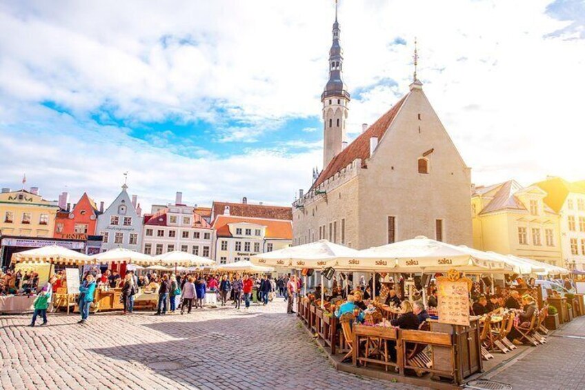 Un mercado al aire libre lleno de gente, comprando y comiendo bajo toldos blancos, con edificios históricos y una imponente torre de iglesia de fondo. 2