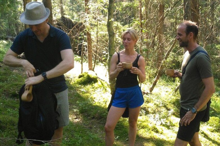 Three people in a forested area, one adjusting a backpack, another holding a cup, and the third observing. 1