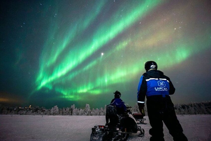 Una persona en un motor para nieve, otra en un trineo, y la aurora boreal al fondo. 2