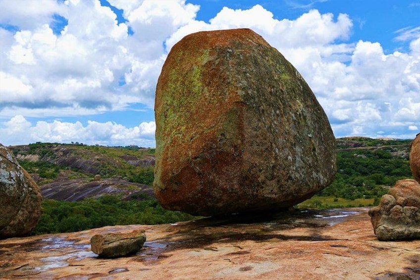 A large rock balanced on a smaller rock, surrounded by a landscape with hills and a partly cloudy sky. 1