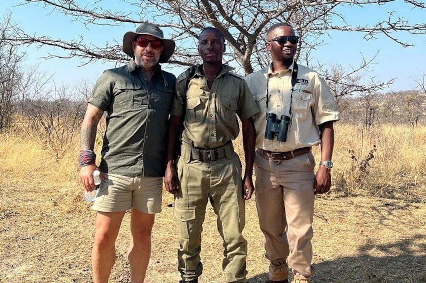 Three individuals in outdoor attire standing in a dry, grassy area with sparse trees in the background. 2