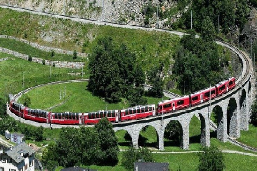 A red train traveling on a bridge with a curved design, surrounded by greenery and a hilly landscape. 4