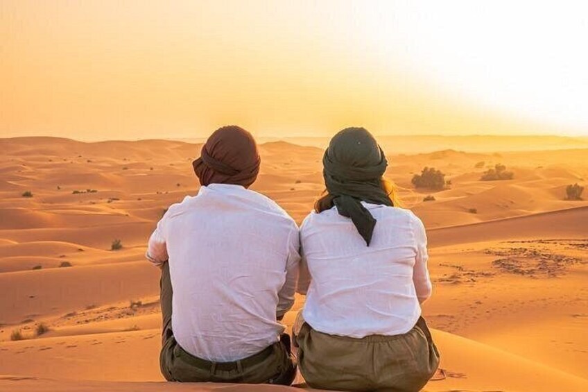 Two people sitting on a sand dune, facing a desert landscape during sunset. 5