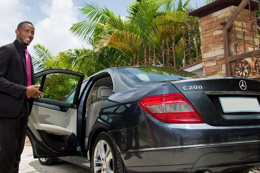 A black Mercedes-Benz C200 parked with its door open, a man in a suit standing beside it, palm trees, and a brick building in the background. 2