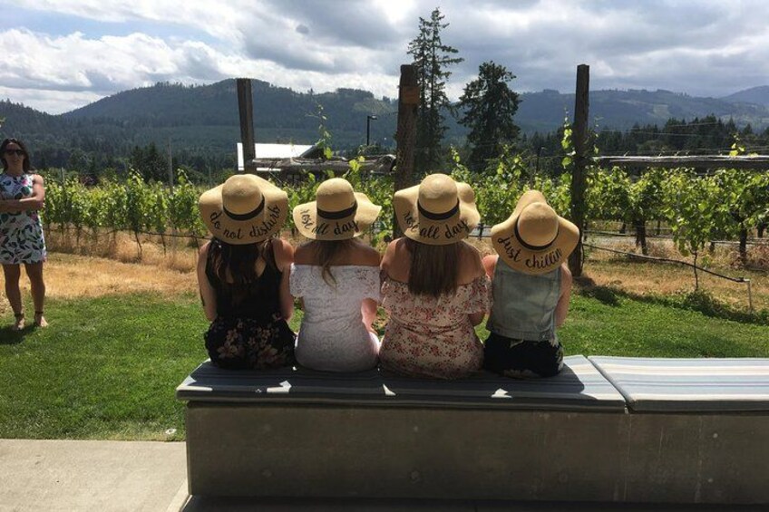 Four people sitting on a bench in a vineyard, wearing hats with phrases on them. 3