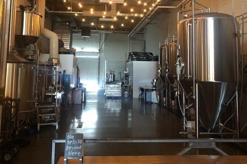 Interior de una cervecería con tanques de acero inoxidable, un cartel de prohibición de bebidas y una pizarra con un mensaje. 5