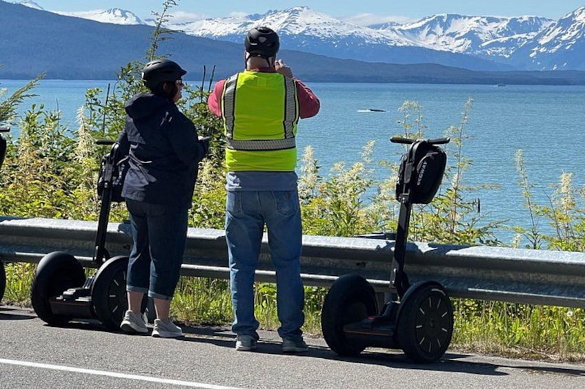 Zwei Personen fahren auf Segways in der Nähe eines Sees mit Bergen im Hintergrund. 2