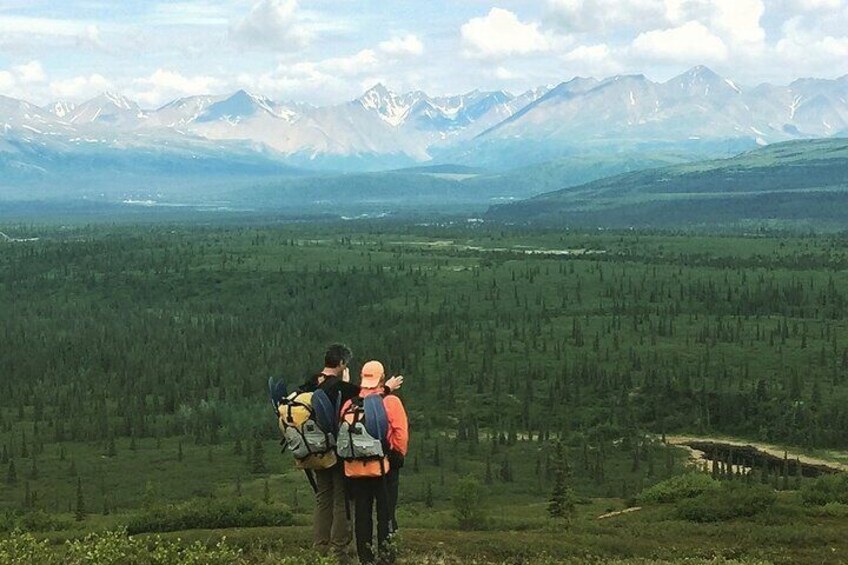 Two hikers with backpacks on a mountain trail overlooking a forested valley and distant mountains. 5