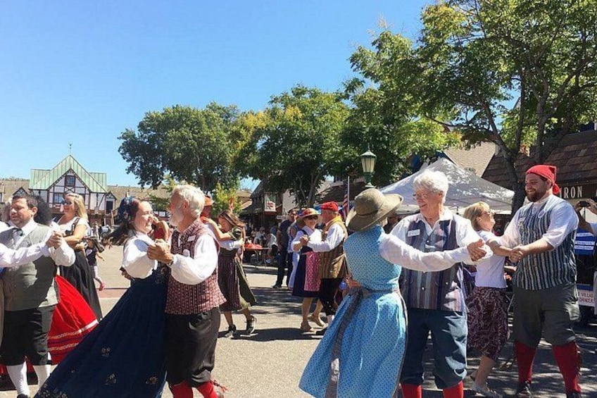 A group of people dancing in traditional costumes in an outdoor setting with trees and buildings in the background. 4