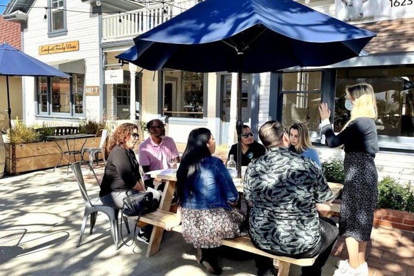 A group of people sitting at an outdoor table with umbrellas, a building with a sign, and a street in the background. 5