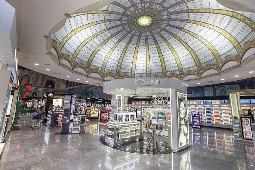 Het interieur van een winkelcentrum met een grote glazen koepel, een Kerastase cosmeticawinkel en diverse winkelkiosken. 2