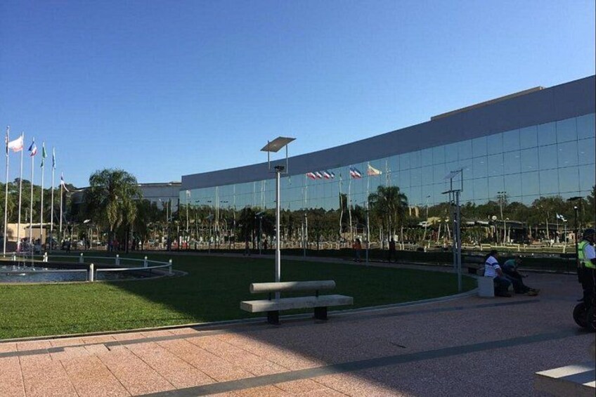 A modern building with a glass facade, a grassy area with a bench, and flags in the background. 4