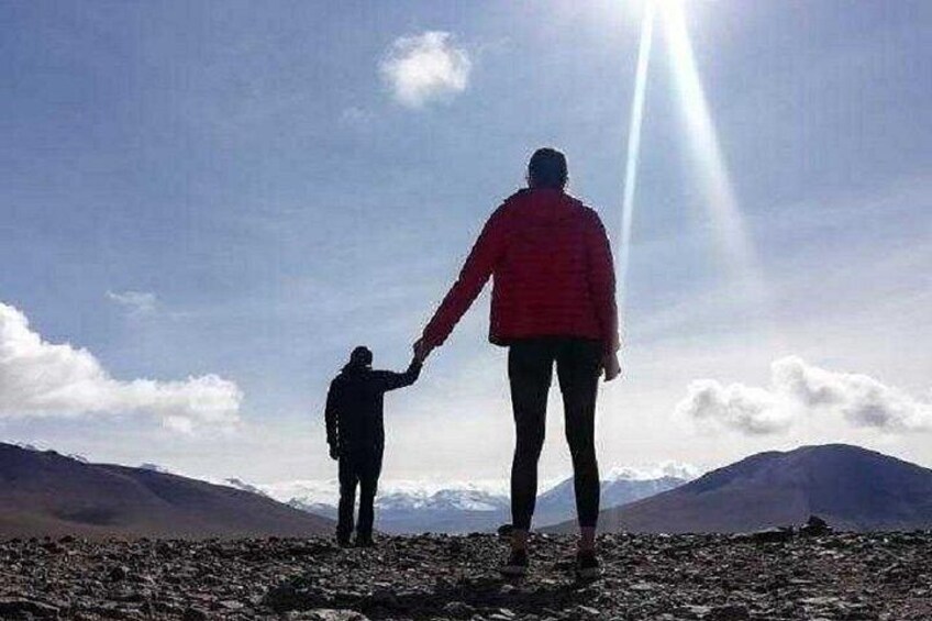 Två personer som håller varandra i handen på en bergstopp med en karg terräng, under en klar himmel och med avlägsna berg i bakgrunden. 2