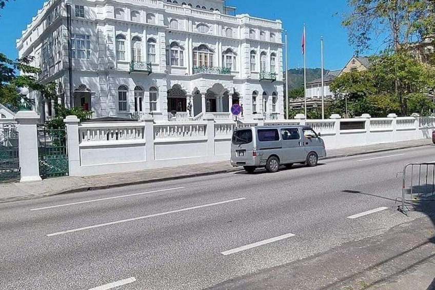 A white building with classical architecture, a road with a white van, and a metal fence. 4