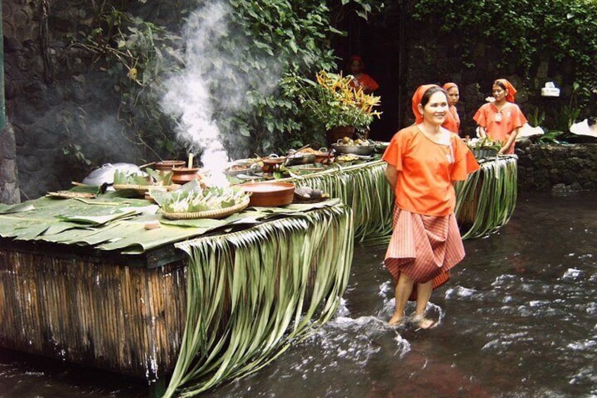 A traditional ceremony with participants in orange attire standing by a water body, surrounded by ritual items and greenery. 1