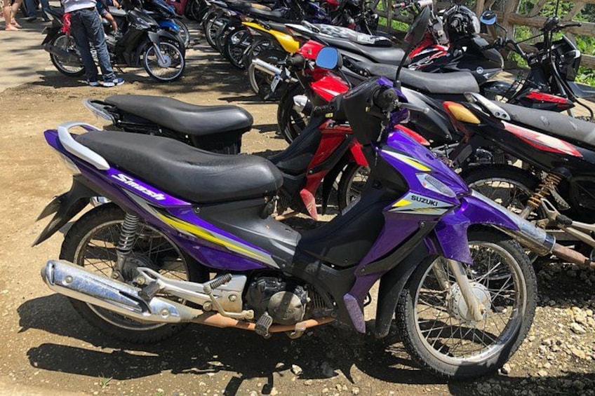 A purple Suzuki motorcycle parked on a dirt surface with other motorcycles in the background. 3