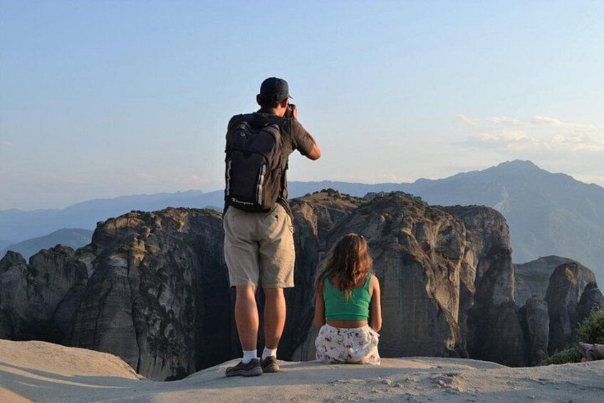 A man with a backpack taking a photo, a woman sitting on a rock, and a mountainous landscape in the background. 5
