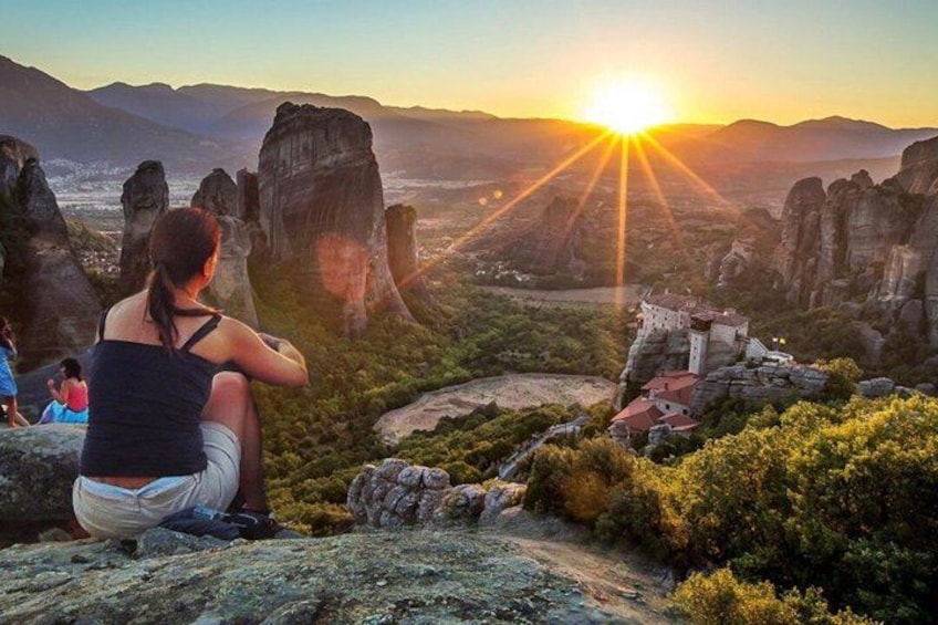 A sunset over a rocky landscape with a person sitting on a rock and a small building in the distance. 4
