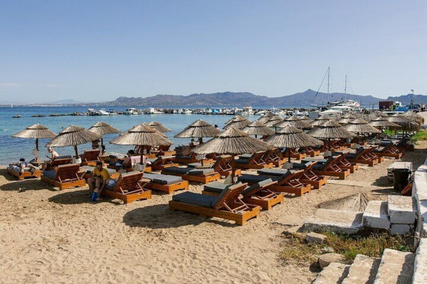A beach with rows of wooden lounge chairs and thatched umbrellas, a clear blue sea, and boats in the background. 5