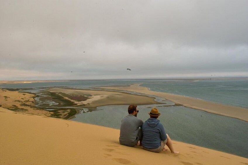 Twee personen zitten op een duin met uitzicht over een kustlandschap, inclusief water en een zandstrand. 1