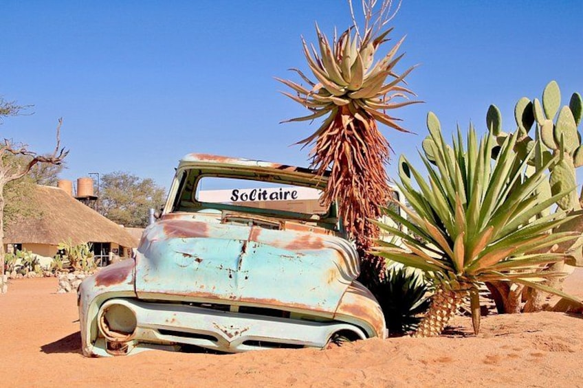 A vintage car with a sign reading 'Solitaire' parked in a desert with cacti and a thatched-roof building in the background. 3