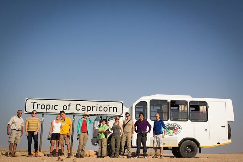 A group of people standing in a desert with a sign that reads 'Tropic of Capricorn' and a white van with 'Tropical Adventure' written on it. 2