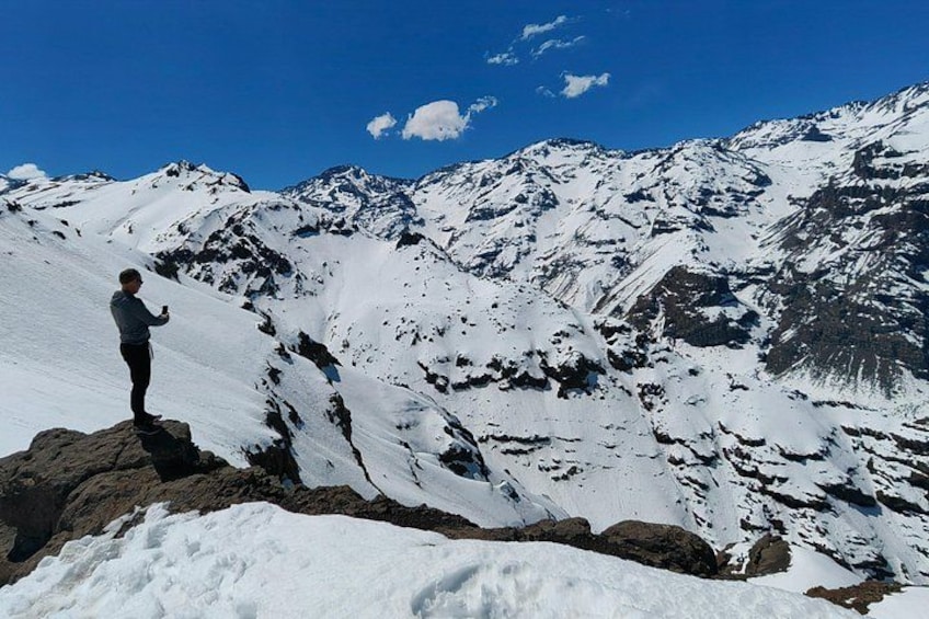 Una persona de pie sobre un afloramiento rocoso, con una cadena montañosa nevada al fondo. 1