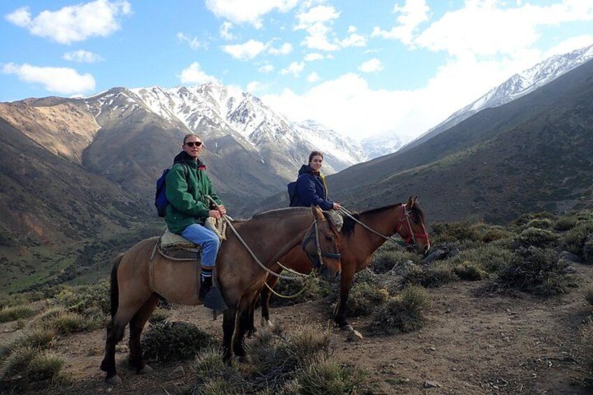 Two people riding horses on a trail with mountains in the background. 1