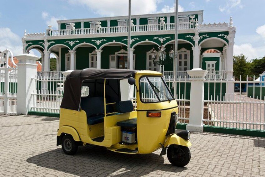 A yellow auto-rickshaw parked in front of a green and white building with ornate architecture. 2