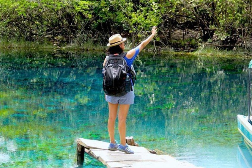 A person standing on a dock extending their arm over water, wearing a hat, shorts, and a backpack. 2