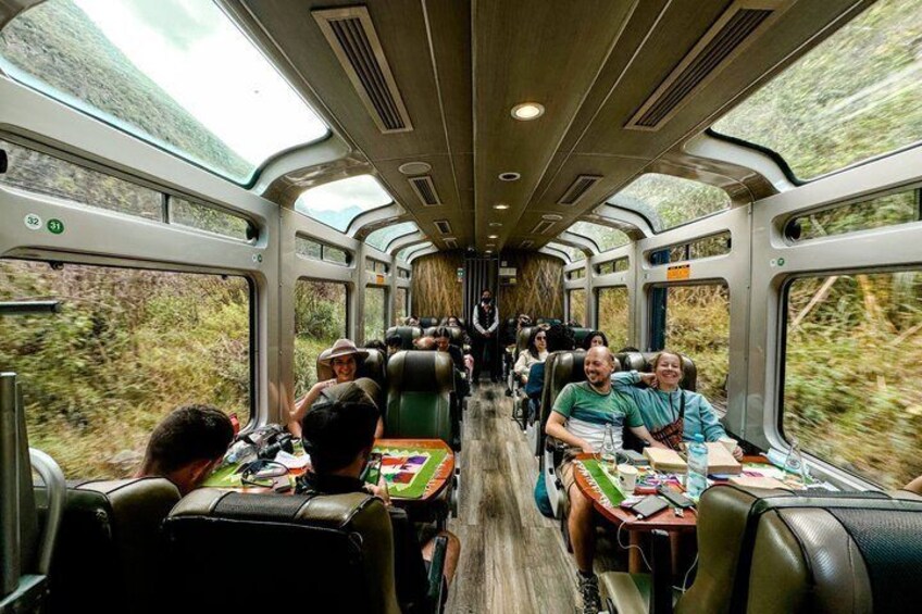 A train interior with passengers seated, windows showing a forested landscape, and overhead luggage compartments. 2