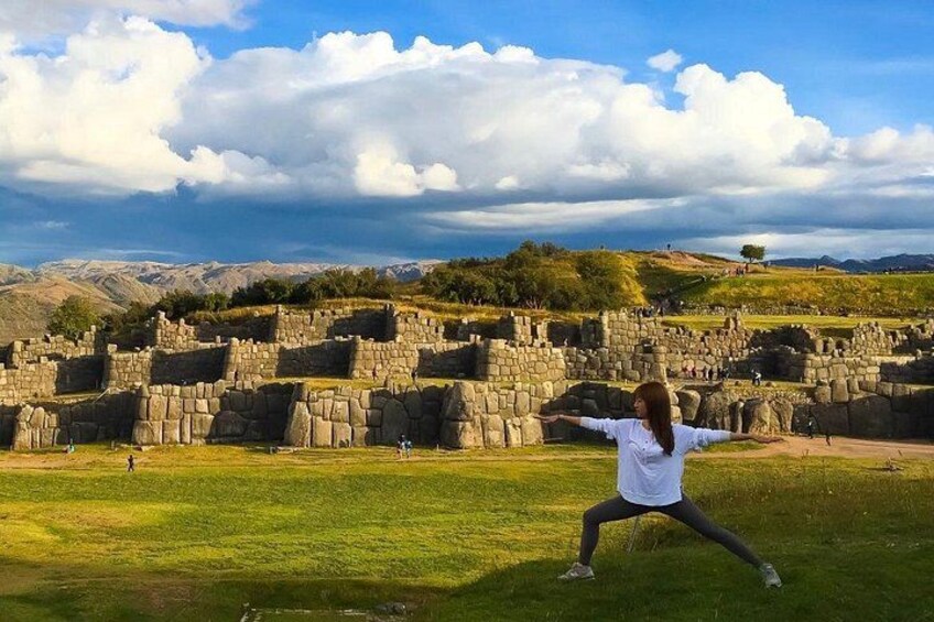 A person in a white shirt and black pants standing with arms outstretched in a grassy area with ancient stone ruins in the background. 2