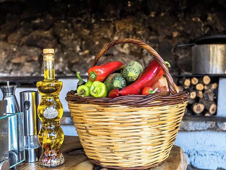 Una cesta de mimbre llena de pimientos verdes y rojos, una botella de aceite y recipientes metálicos sobre una superficie de madera, con un muro de piedra y una olla al fondo. 4