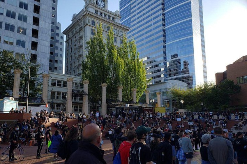 A crowded public square with tall buildings and trees. 2