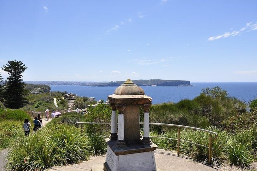 Camino costero con un monumento de piedra, vegetación y una vista distante del mar y la tierra. 2