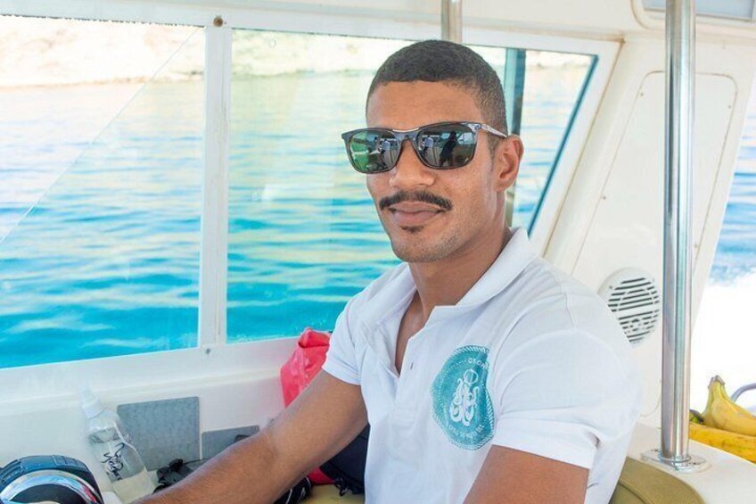 Una persona con camisa blanca y un logo, manejando un bote con vista a aguas azules a través de una ventana. 3