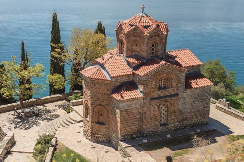 A stone church with a red-tiled roof, surrounded by trees and a body of water. 2