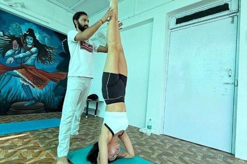 A person performing a headstand with assistance, in a room with a mural of a deity and a closed door. 4