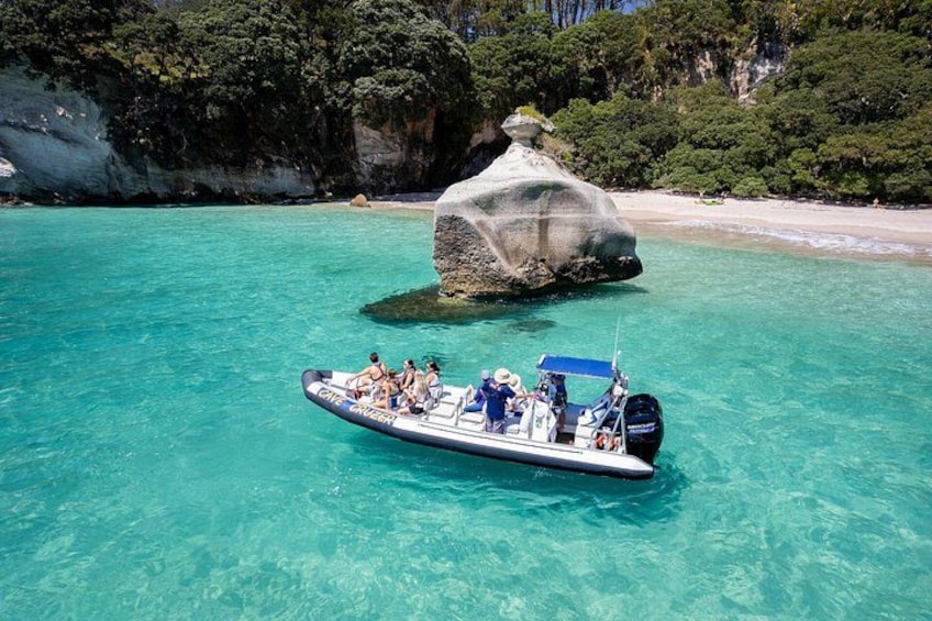 A boat with people on a clear turquoise sea, a large rock formation, and a beach with greenery in the background. 1