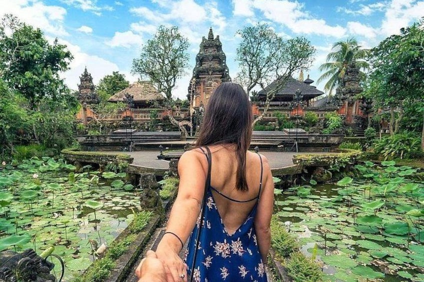 A woman in a blue dress standing by a pond with lotus flowers, with temple structures in the background. 1