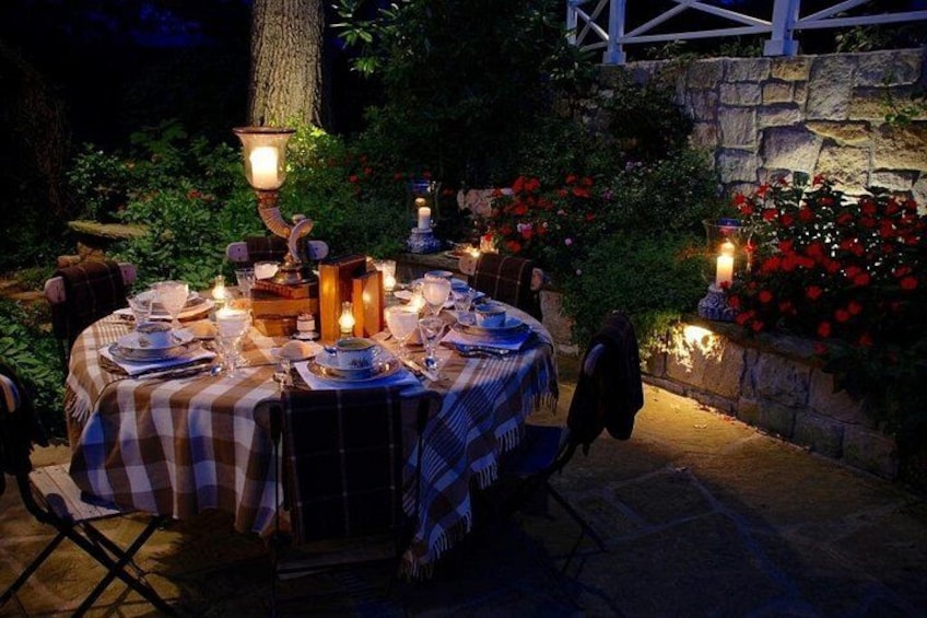 An outdoor dining area with a table set for a meal, surrounded by stone walls and flowering plants. 3