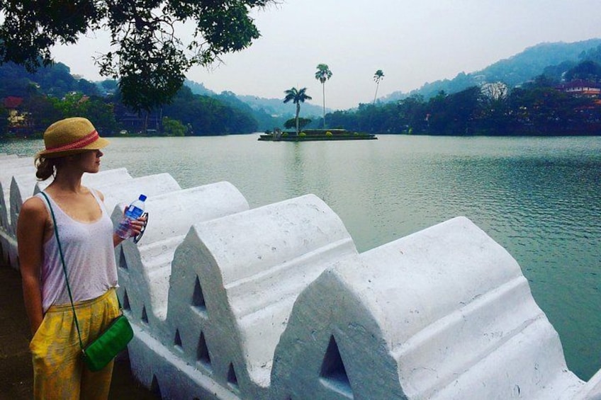 A person standing by a calm lake with a white stone barrier, surrounded by greenery and hills. 1