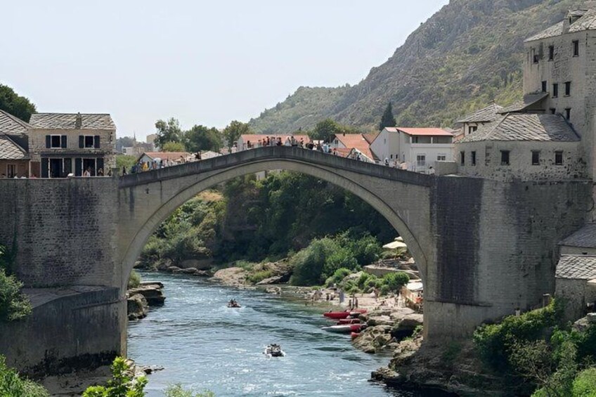 A stone bridge over a river with boats, buildings, and a hilly landscape. 5