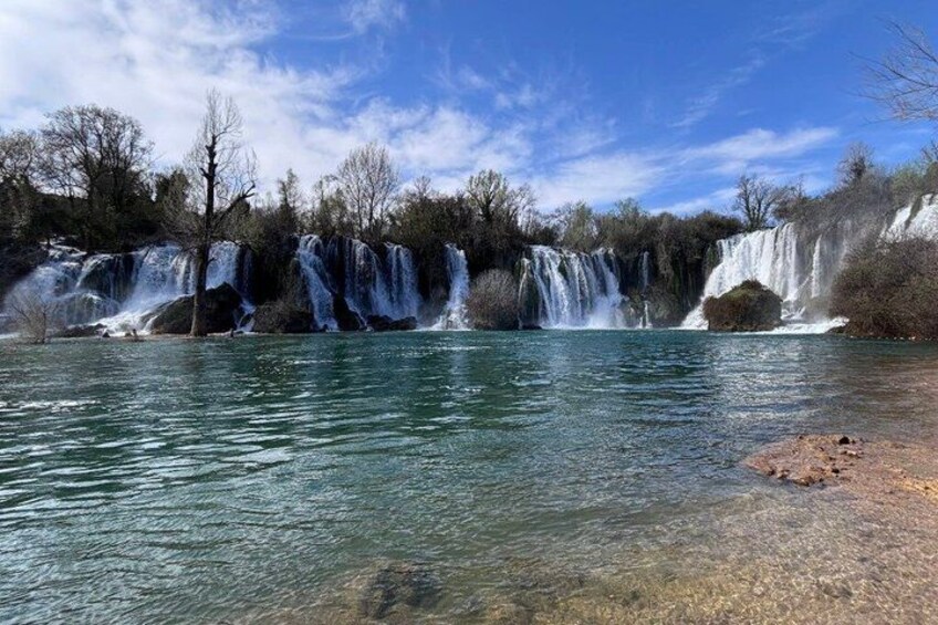A waterfall cascading over rocks into a clear body of water, surrounded by trees and a clear sky. 4